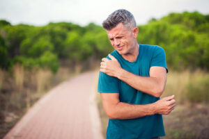 Man in blue T-shirt on jogging path clutching his painful shoulder